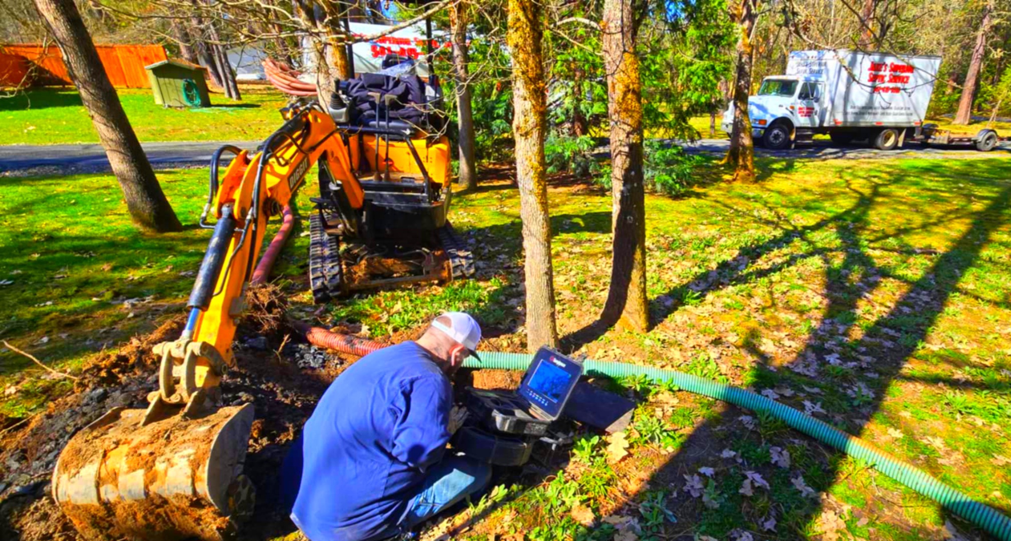 Septic technician performing a camera inspection at a residential property, using equipment beside an excavated septic tank with a service truck and mini excavator in the background.