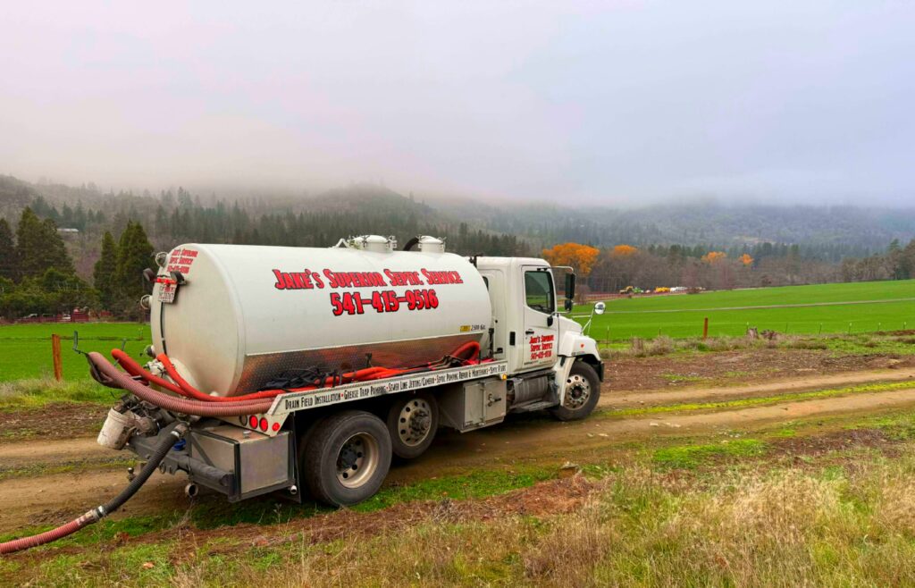Septic pumping truck from Jake’s Superior Septic Service parked on a rural dirt road, servicing a property with green fields and forested hills in the background.